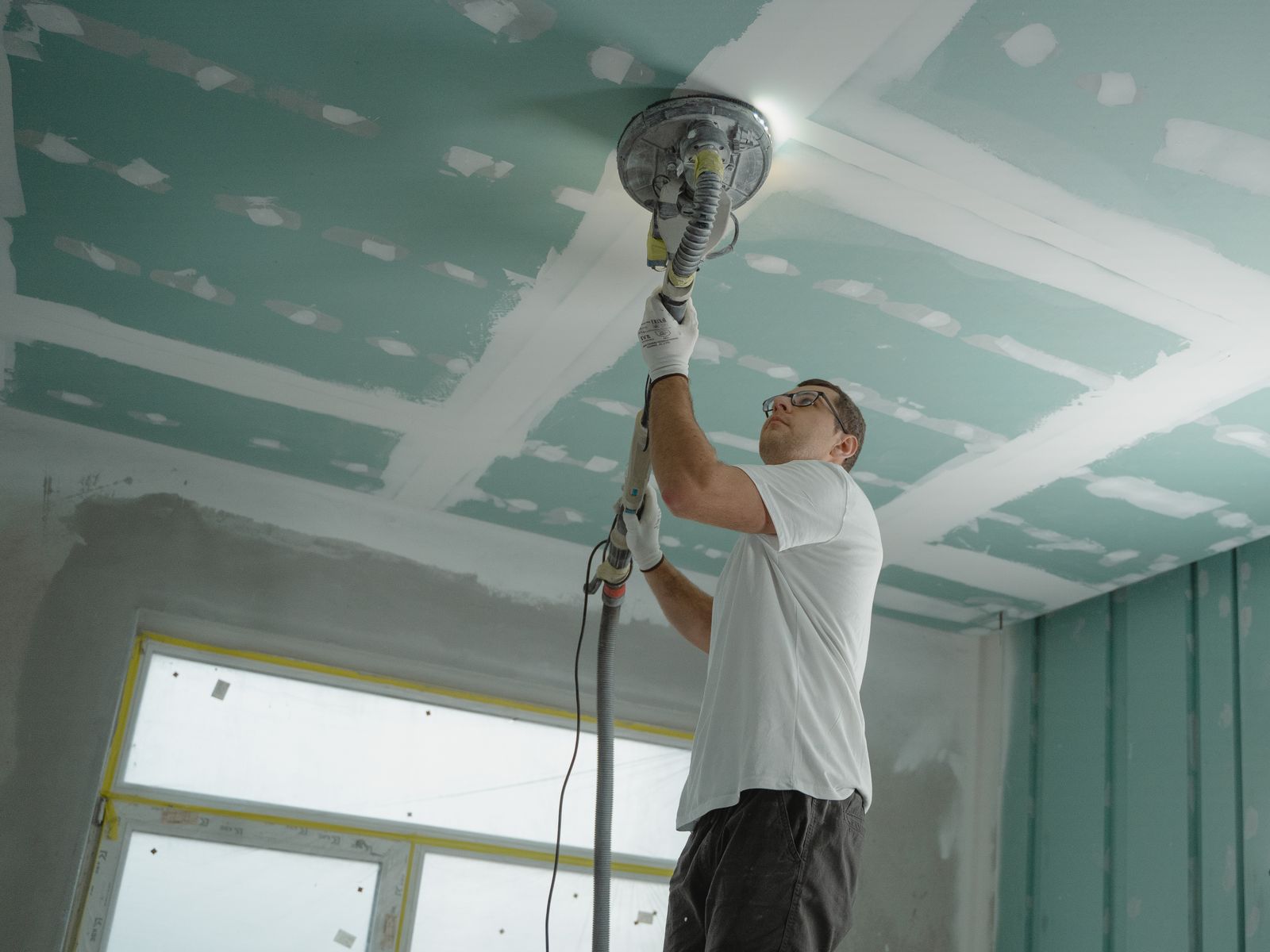 Contractor sanding a drywall ceiling smooth with a dust-controlled orbital sander — the refinishing stage of popcorn ceiling removal